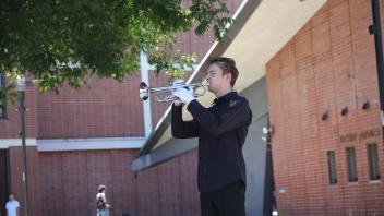Young man plays trumpet outside by a red-brick building under a leafy tree