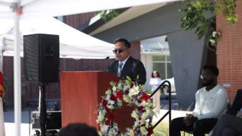 A man in a suit and sunglasses speaks at a podium beside a floral wreath, with seated guests and onlookers behind him, during the UC Davis Memorial Day Ceremony 2025