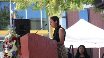 A woman with braided hair stands at the podium reading from a paper while another speaker waits behind her, during the UC Davis Memorial Day Ceremony 2025