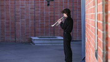 A young man dressed in black stands alone playing a trumpet in a quiet brick courtyard