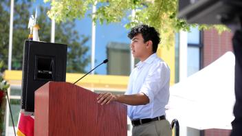 A young man stands at a podium under leafy trees, delivering a speech