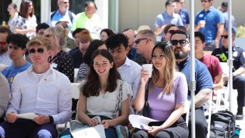 Attendees of various ages sit in rows under a canopy, holding programs and chatting warmly