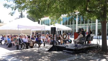 Dozens of attendees sit under white canopies listening to speakers on stage