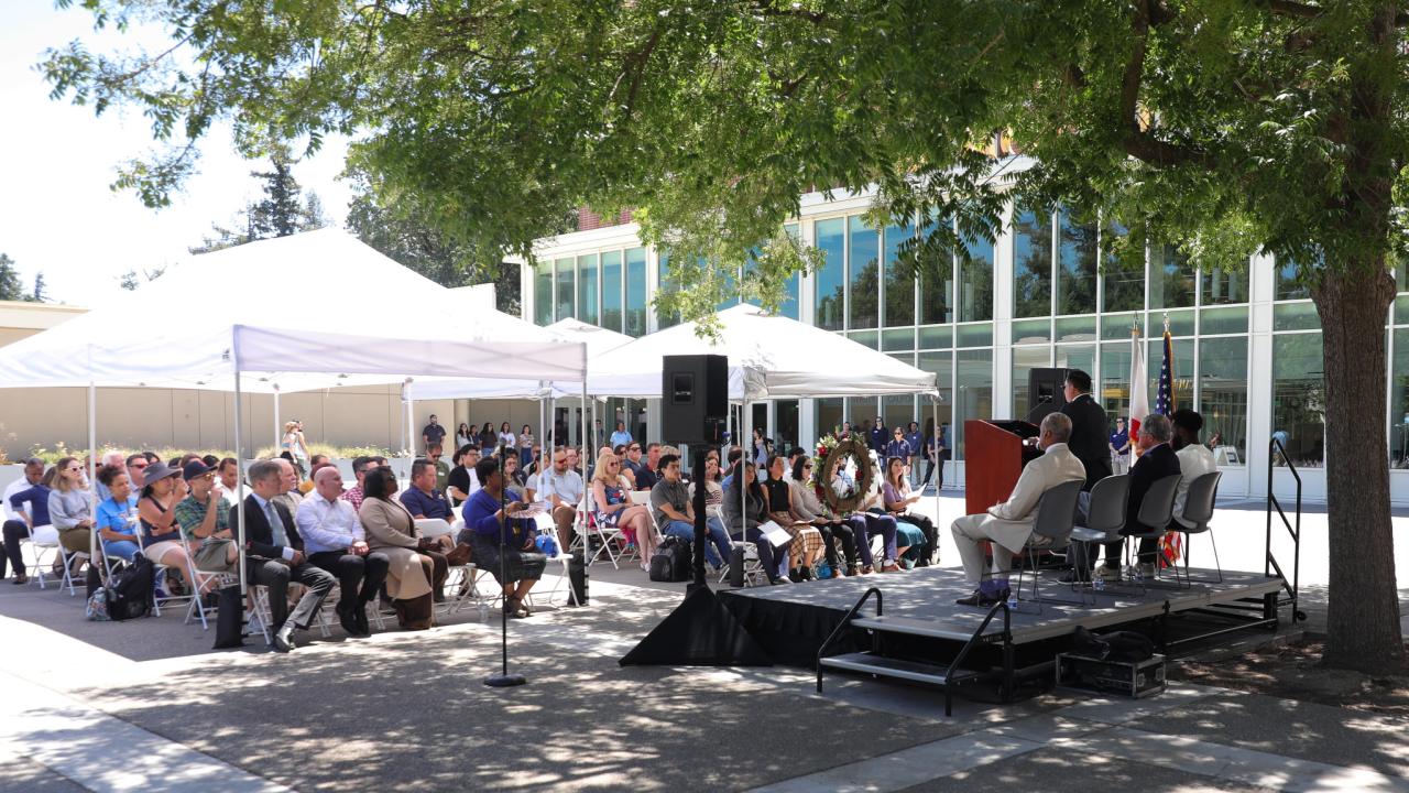 Dozens of attendees sit under white canopies listening to speakers on stage