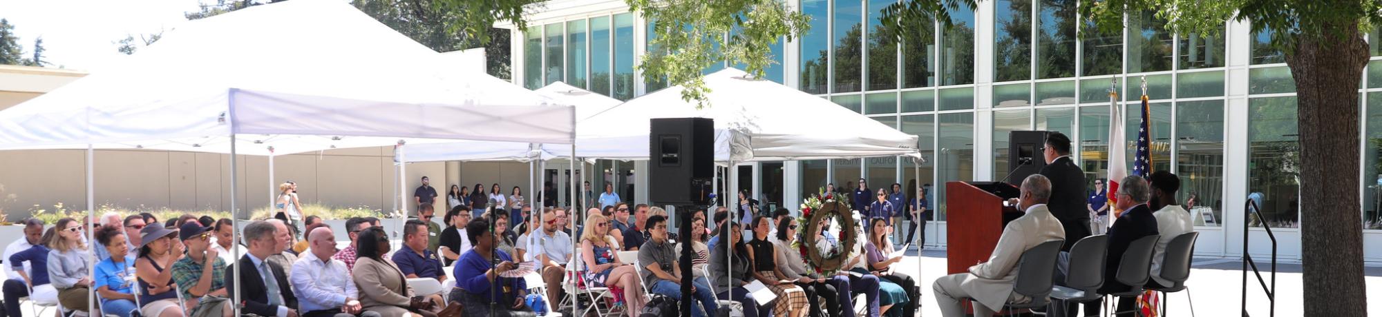 Dozens of attendees sit under white canopies listening to speakers on stage